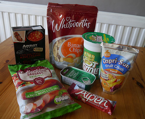 Food on a table - instant soup, banana chips, pot noodle, drink ceral bar, tinned fish, sausage